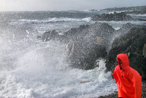 Picture of waves crashing over rocks, spray coming over. Also a boy in a red waterproof jacket