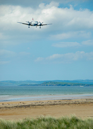 Picture of a plane coming in over a bay with a beach to land on an airport near the shore