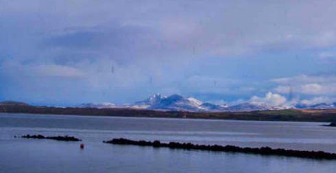 Picture of snow covered mountains seen in the distance across a sea loch and some lower hills