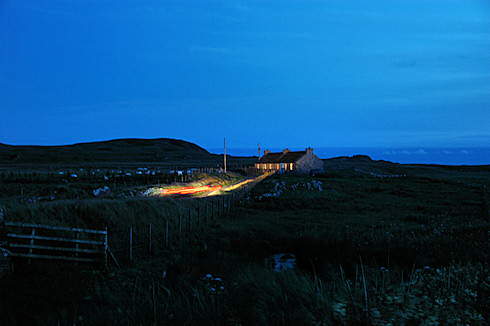 Picture of a cottage at a single track road at night, a car approaching with the lights leaving their marks on the picture