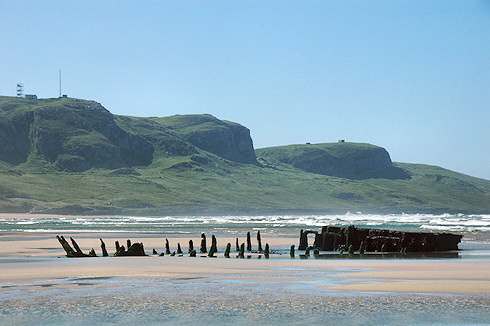 Picture of a wreck on a beach, high cliffs in the background, all under a brilliant blue sky