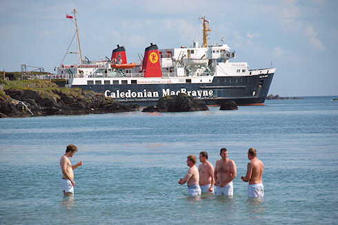 Picture of a group of men standing in the water off a beach, a Calmac ferry in the background