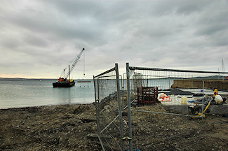 Picture of a construction fence around a water treatment outlet, a barge with a crane in the background