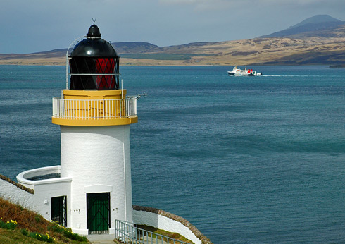 Picture of a lighthouse on a hillside above a sound, a Calmac ferry passing it