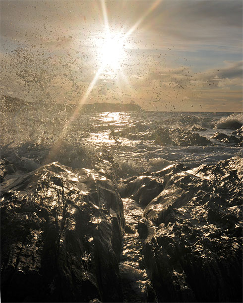 Picture of a wave splashing over rocks in a bay with the low sun in the background