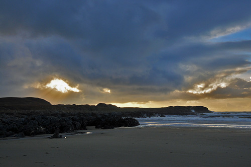 Picture of the sun breaking through some clouds over the cliffs of a sandy bay