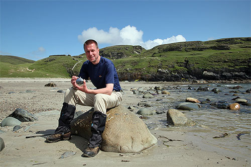 Picture of a man sitting on a rock on a beach with a Laphroaig hip flask in his hands