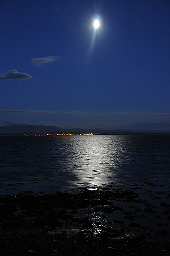 Picture of the Moon over a sea loch, reflecting in the loch. The lights of a village in the distance